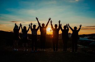 Fotografische Darstellung mehrerer Personen, die nebeneinander  vor einem Sonnenuntergang stehen. Die Personen fassen sich an die Hände und strecken diese in die Luft. Gesichter, Geschlecht und Alter der Personen sind nicht zu erkennen.